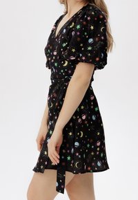 Czarna sukienka kopertowa z bufkami, ozdobiona kolorowymi motywami niebiańskimi, takimi jak gwiazdy, księżyce i planety. Materiał o fakturze, wiązana w talii.