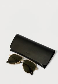 Óculos de sol com armação dourada e lentes escuras, apresentando pontas de haste pretas, colocados ao lado de uma elegante caixa de couro preto com uma textura suave.