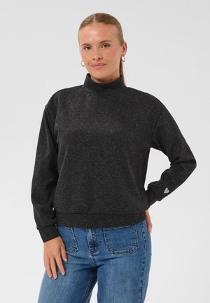 Femme aux cheveux châtain clair en queue de cheval, portant un haut gris foncé à manches longues et un jean bleu, debout avec une main sur la hanche devant un fond uni.