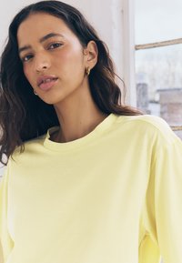 Hellgelbes Langarmoberteil mit rundem Ausschnitt und lockerem Sitz. Glatte Stoffstruktur, minimale Nähte, keine Muster oder Akzente.