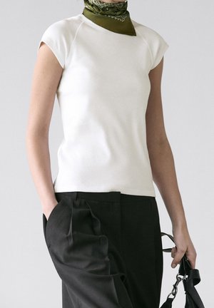 Personne portant un haut blanc à manches courtes, un pantalon noir plissé, une écharpe verte à motifs autour du cou, tenant la sangle d'un sac à main noir.