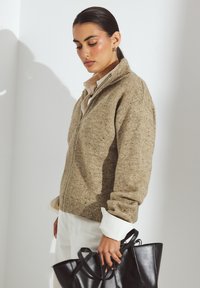 Sadrāžots džemperis ar rāvējslēdzēju gaisā brūnā krāsā ar punktainām svītrām, apakšā valkājot krēmkrāsas krekliņu un melnu somu. Mīksta tekstūra, brīva piegriezuma.