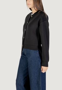 Felpa nera con scollatura rotonda e chiusura con bottoni sul davanti. Presenta maniche lunghe e polsini a coste. Indossata con jeans blu larghi.