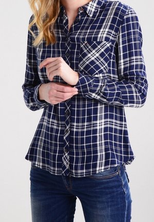 Kvinde justerer ærmet på blå- og hvidternet skjorte med knapper, båret med blå jeans mod ensfarvet baggrund.