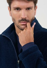 Chaqueta de forro polar en color navy con un alto cuello, cierre frontal con cremallera y textura acanalada. En primer plano se muestran manos tocando el cuello, revelando una tela interior de contraste.