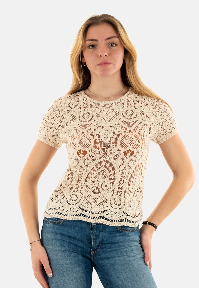 Top de crochet de color crema con un intrincado patrón floral, cuello redondo y textura semitransparente, combinado con jeans de mezclilla azules.