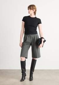 Svart teksturert topp med korte ermer, olivengrønne vide shorts og knelang svarte støvler. Holder en svart clutch, står på en nøytral gulv.