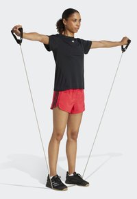 T-shirt de sport noire, shorts rouges avec des rayures noires, chaussures de sport noires. Tenant des bandes de résistance, bras étendus sur les côtés.