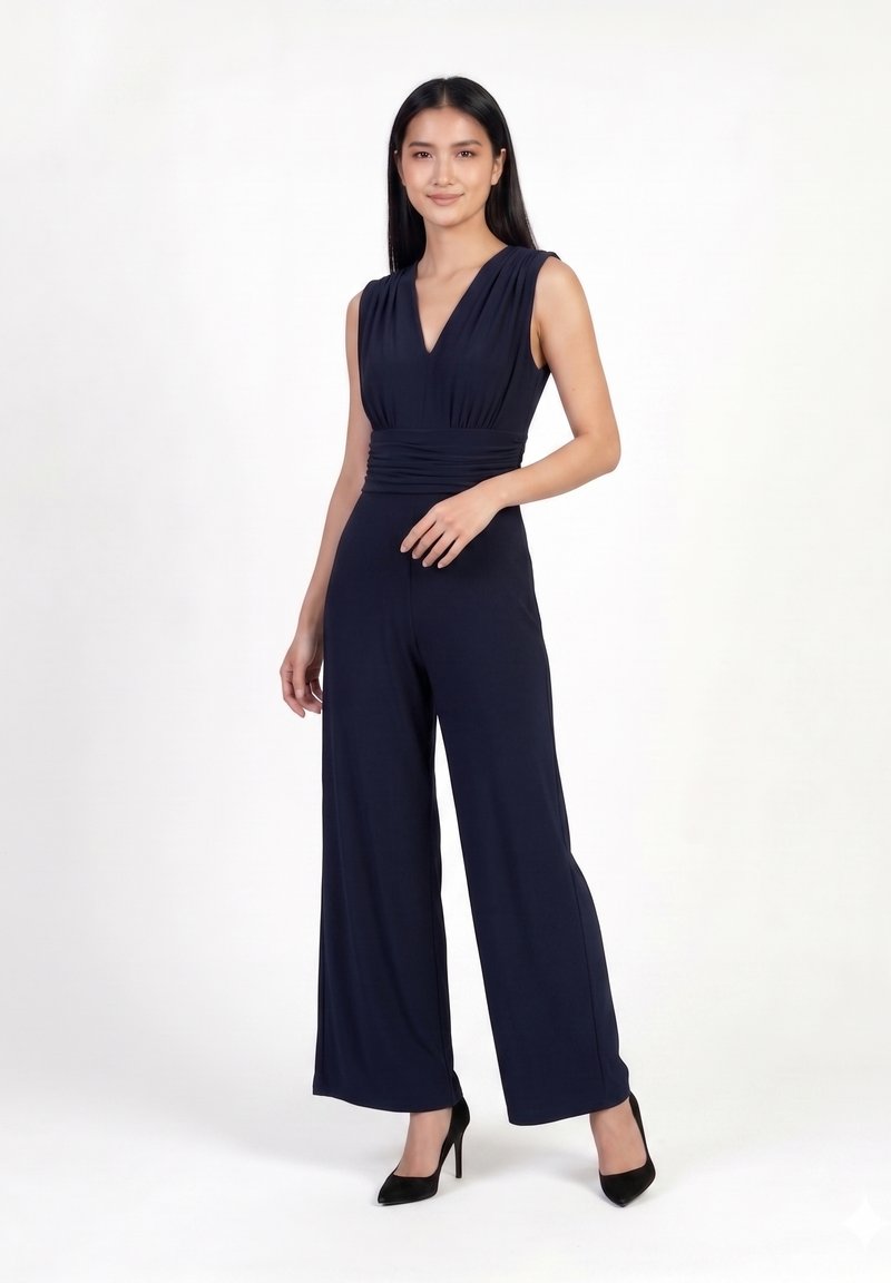 Femme debout dans une combinaison sans manches bleu marine à jambes larges et col en V, portant des talons noirs sur fond blanc.