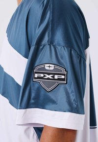 Maglia sportiva a maniche corte in blu e bianco con una patch logo a scudo "PXP" sulla manica superiore.