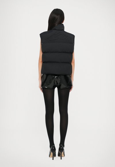 Sort puffer vest med høj krave og stikninger, båret over skinnende sorte shorts. Sorte strømpebukser og højhælede sko fuldender looket.