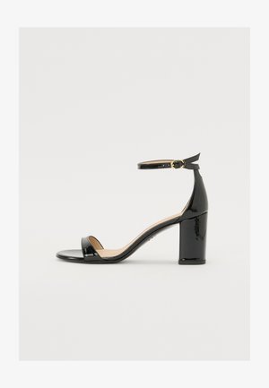Black patent leather sandal with a wide heel, open toe, and ankle strap featuring a gold buckle. Smooth texture and sleek design.