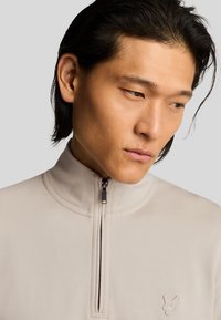 Lichtbeige rits-pullover gemaakt van zachte stof, met een hoge kraag, subtiele stiksels en een logo-opdruk op de borst.