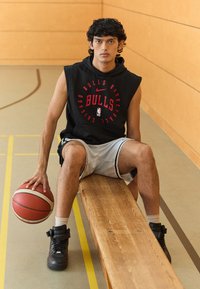 Črn brezrokavnik sredi z rdečim napisom "BULLS", sivi kratki hlače in črne superge. Sedi na leseni klopi in drži košarkarsko žogo.