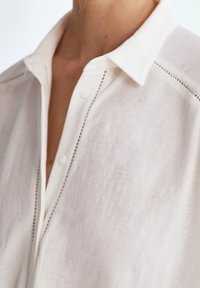 Weißes Hemd mit Kragen und Knopfleiste, mit feinen Stickdetails entlang der Nähte, aus einem leichten, strukturierten Stoff gefertigt.