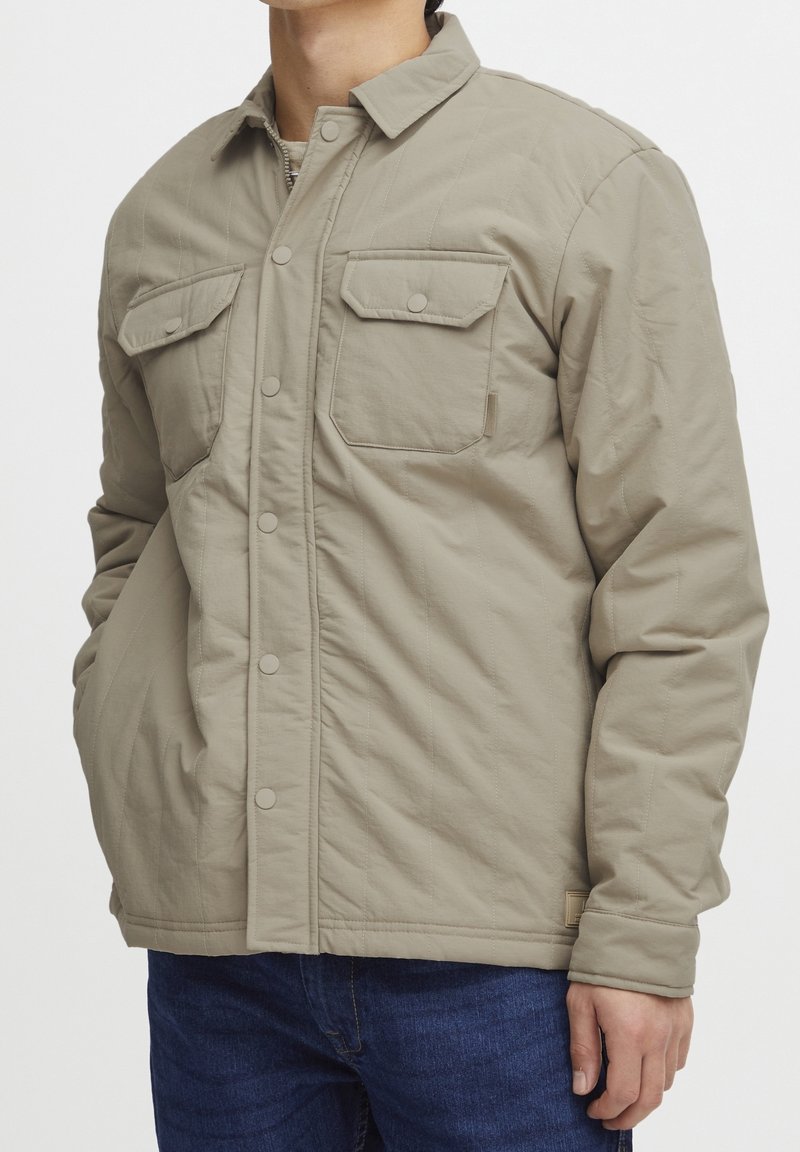Beige quiltet jakke med trykknaplukning, to brystlommer med klap, båret af person i blå jeans med højre hånd i lommen.