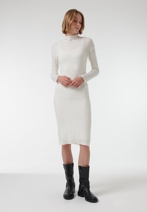 Robe en tricot blanc à manches longues, avec une texture côtelée et une silhouette ajustée. Associée à des bottines noires avec des boucles.