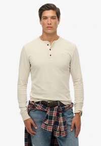 Camisa henley beige de manga larga con tres botones oscuros, combinada con jeans azules y una camisa de cuadros rojos atada a la cintura. Cinturón marrón visible.