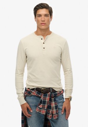 Beige Langarm-Henley-Shirt mit drei dunklen Knöpfen, kombiniert mit blauen Jeans und einem rotkarieren Hemd, das um die Taille gebunden ist. Brauner Gürtel sichtbar.