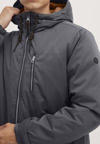 Gray insulated jacket with a hood, featuring zippered front pocket and contrasting orange lining. Includes adjustable drawstrings and logo patch.