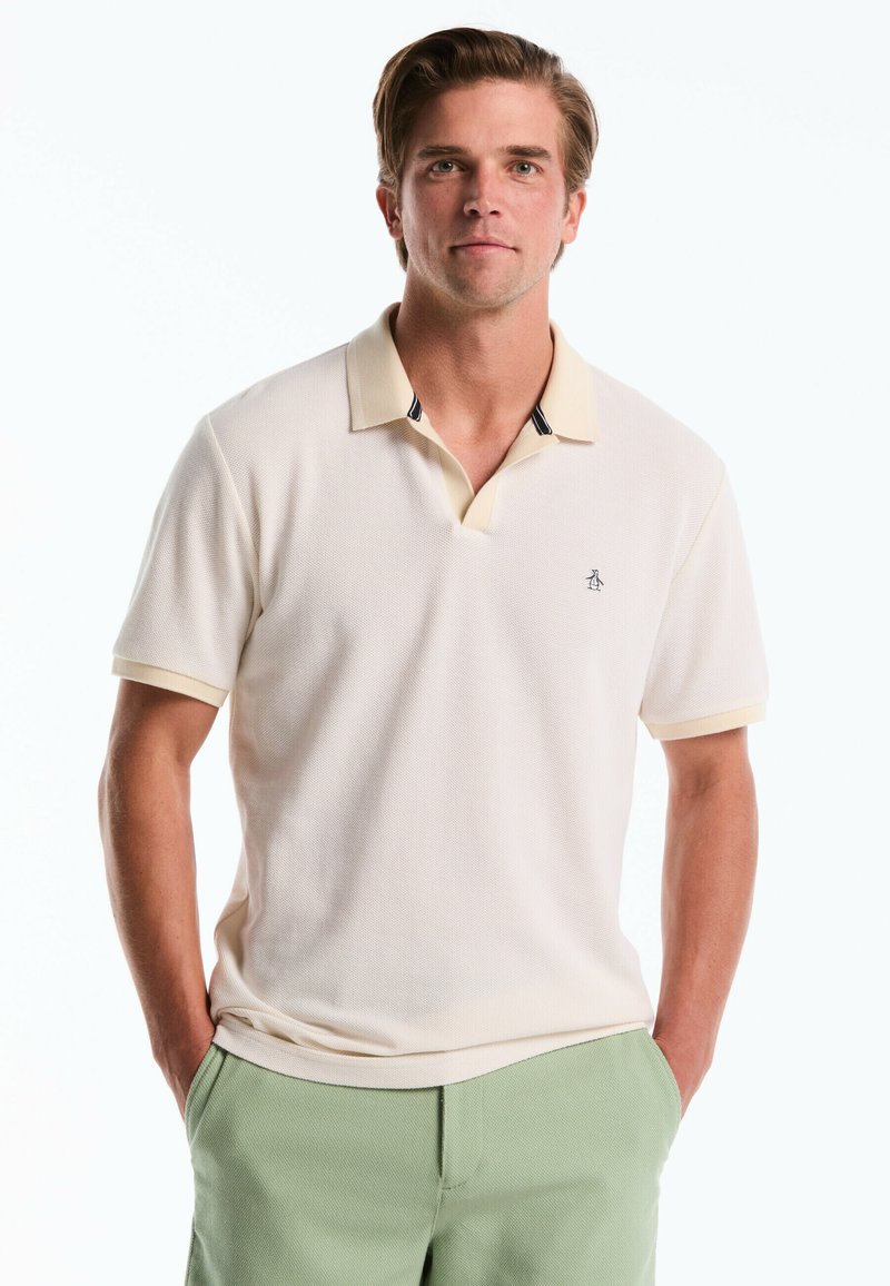 Crèmekleurige polo met een gestructureerde stof, voorzien van een v-hals kraag met lichtgele bies en donkere accenten. Klein logo op de borst.