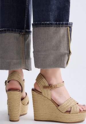 Pies con sandalias de cuña beige tejidas con tiras en el tobillo, combinadas con vaqueros oscuros remangados en los puños.