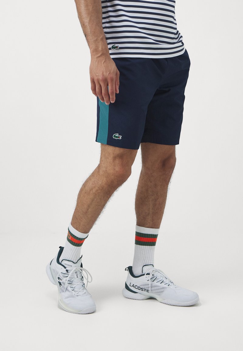 Shorts bleu marine avec un panneau latéral teal, arborant un logo Lacoste vert. Portés avec des baskets blanches et des chaussettes de couleur rayée.