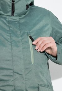 Giacca verde a spina di pesce con colletto alto, dotata di tasca sul petto con zip e texture liscia, realizzata in un materiale leggero.