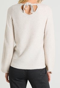 Cremefarvet sweater med lange ærmer, en struktureret overflade og en hjerteformet udsparing bagpå med en bundet bånddetalje.