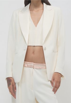 Frau trägt einen cremefarbenen Blazer, ein kurzes Top und passende Hosen mit rosa Bund, zeigt ein Rosentattoo auf ihrer linken Hand.