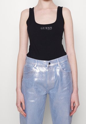 Osoba nosząca czarny top bez rękawów z logo „GUESS” oraz błyszczące jasnoniebieskie dżinsy z wysokim stanem, stojąca na jednolitym tle.