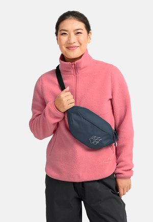 Vrouw draagt een roze fleecejack en zwarte broek, houdt de band vast van een donkerblauwe crossbodytas met een pootafdruklogo.
