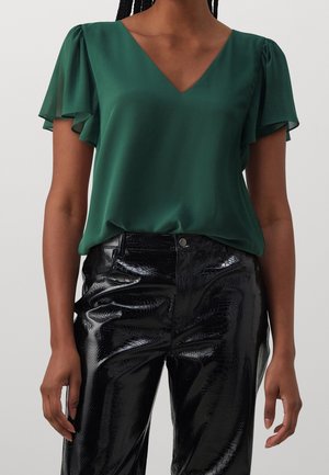 Femme portant un chemisier vert foncé à col en V avec des manches volantées et un pantalon noir brillant texturé taille haute.