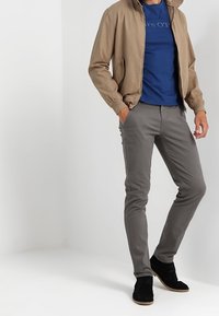 Tan bomberjacka, blå t-shirt med logotyp, grå slim-fit byxor, svarta ankelskor. Slät tyg, avslappnad design, enkel färgsättning.