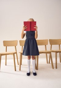 Et barn i en mørk denimkjole med sløjfe, hvide knæstrømper og sorte sko holder en rød bog og står foran træstole.