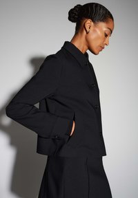 Zwarte cropped jas met een gestructureerde kraag en knoopsluiting. Voorzien van zijzakken en een textuurstof, gedragen bij een bijpassende outfit.