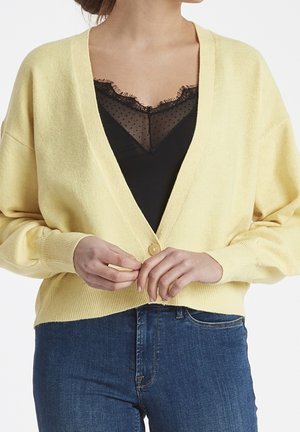 Gul kofta med v-ringning, som har knäppning fram och ribbade ärmslut. Under är en svart topp med spetsdetaljer och prickar.