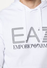 Bílá mikina s kapucí, která má velké černé logo ve stylu síťoviny se nápisem "EA7 EMPORIO ARMANI." Včetně stahovacích šňůrek a standardního střihu s měkkou texturou.