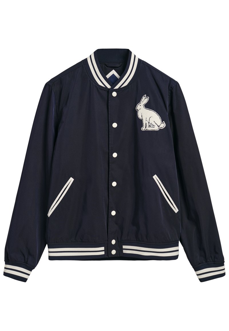 Marineblau bomberjacke mit weißen Streifen an den Bündchen und am Kragen. Hat einen weißen gestickten Hasen auf der linken Brust und zwei Seitentaschen.