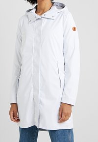 Cappotto impermeabile grigio chiaro con cappuccio, chiusura frontale a bottoni a scatto, tasche laterali e patch con logo sul braccio sinistro. Tessuto liscio e design di lunghezza media.