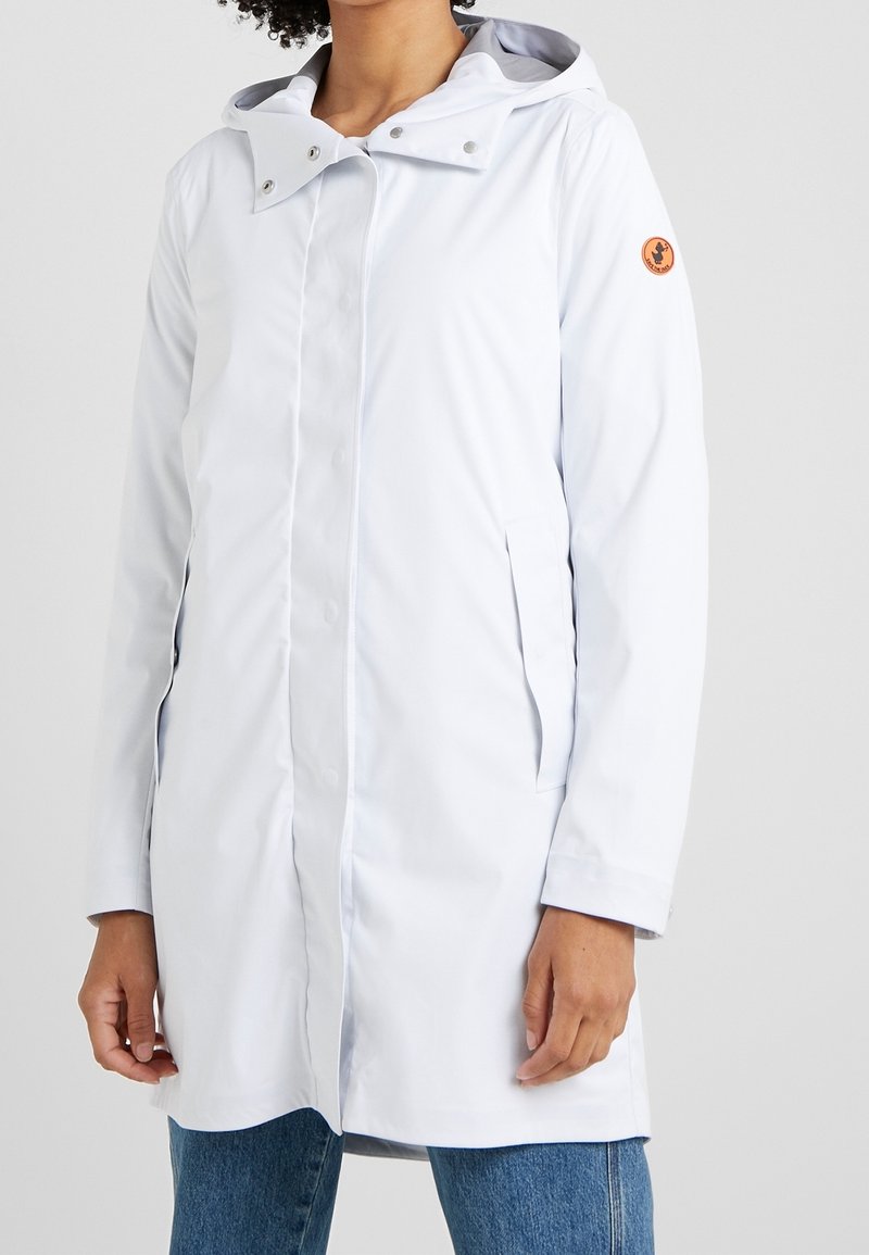 Manteau imperméable gris clair avec capuche, fermeture à boutons-pression, poches latérales et écusson logo sur le bras gauche. Texture lisse et coupe mi-longue.