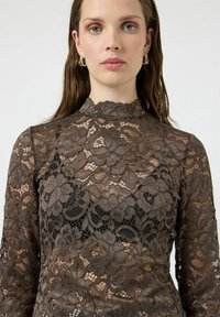 Blondebluse med lange ermer i jordnær brunfarge, med blomsterdesign og høy hals. Gjennomsiktig design med svart stoff under.