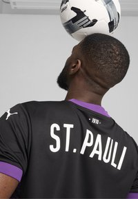 Camiseta negra con el texto "ST. PAULI" en blanco, acentos de color púrpura en el cuello y el logo de Puma en la manga. Un balón de fútbol está equilibrado en la cabeza.