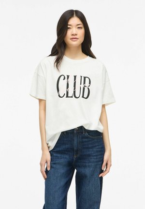Jeune femme aux longs cheveux foncés portant un t-shirt blanc oversize avec le texte "CLUB" et un jean bleu foncé, debout devant un fond blanc uni.