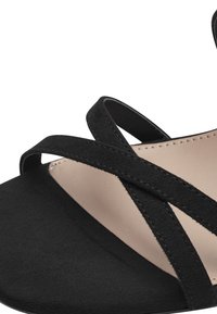 Mustat mokkasandaalit, joissa on ristiin menevät hihnat, litteä pohja ja sileä beige sisäpohja. Minimalistinen muotoilu, jossa on teksturoitu päällisosa.