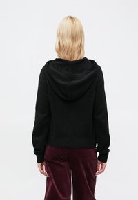 Μαύρο φούτερ με υφή υφάσματος, ριμπ μανσέτες και τελείωμα. Διαθέτει άνετη εφαρμογή και μεγάλο κουκούλα. Φορεμένο με μπορντό παντελόνι κοτλέ.