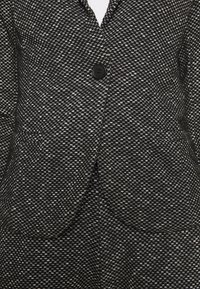 Blazer texturé tissé noir et blanc avec un seul bouton noir et deux poches avant, porté sur un top blanc.