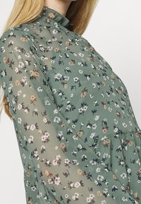 Blusa verde fluida en tejido transparente con un patrón floral en blanco, marrón y azul oscuro, con cuello rizado y mangas largas.