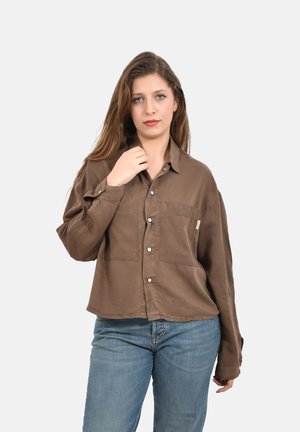 Giovane donna con capelli lunghi che indossa una camicia marrone con bottoni e jeans blu, in piedi davanti a uno sfondo chiaro e uniforme.