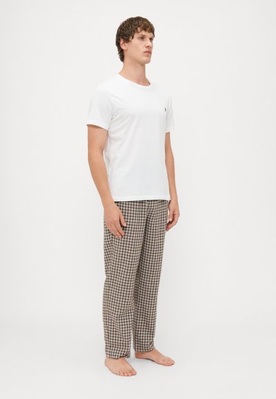 Witte katoenen t-shirt met korte mouwen, gecombineerd met bruin-en-crème geruite pyjamabroek, staand op blote voeten tegen een effen achtergrond.